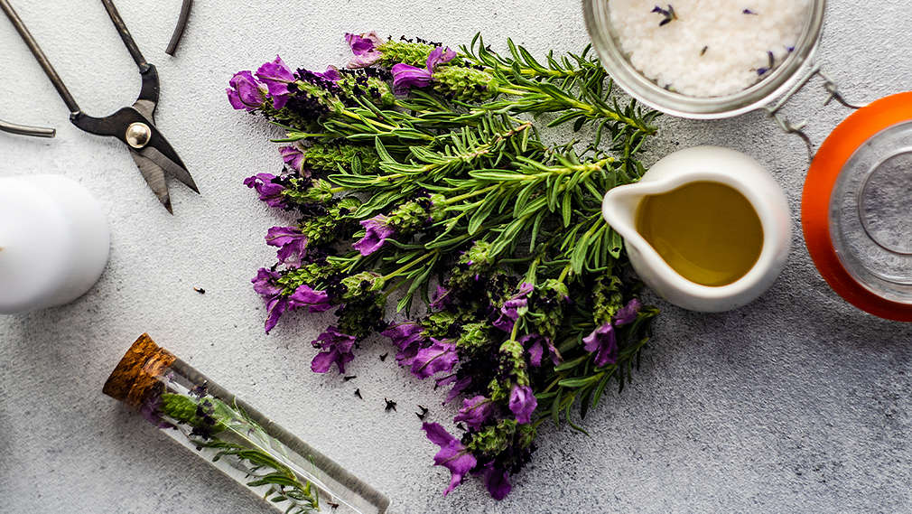 Frischer Lavendel liegt neben einem Bügelglas mit selbstgemachtem Badezusatz mit Lavendel sowie einem Fläschchen Lavendelöl, weißen Kerzen und einer Schere auf einem hellgrauen Grund