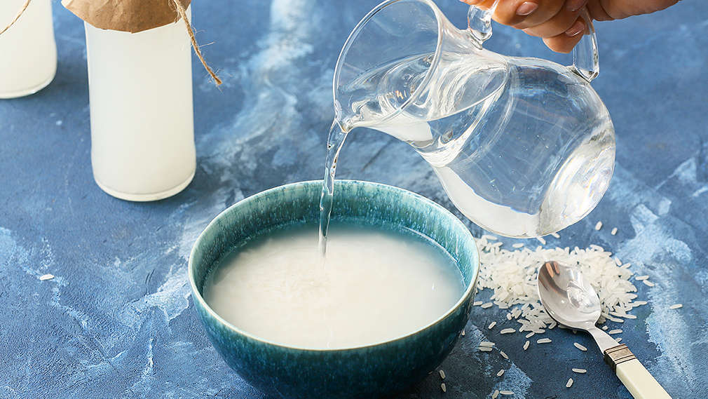 Blauer Untergrund, darauf milchig-weiße Plastikbehälter, ein Löffel mit weißem Griff, Reiskörner und eine türkise Schale mit Reiswasser, in die gerade Wasser aus einem Krug gegossen wird, Hand im Anschnitt.