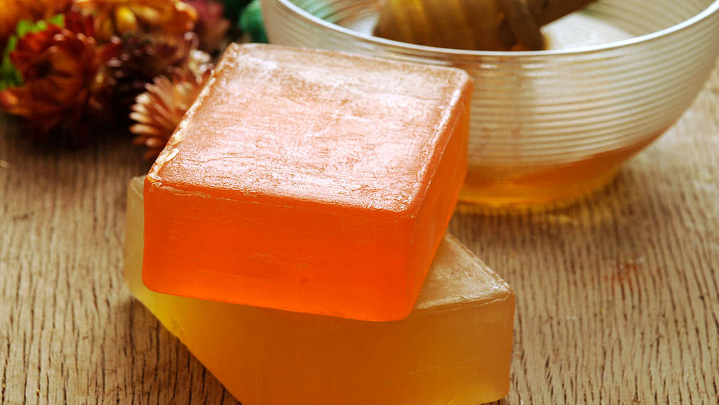 Zwei Stück handgemachte Seife – oben in oranger, unten in gelber Farbe, die übereinander gestapelt auf Untergrund aus Holz liegen. Im Hintergrund ist eine Glasschale mit Honig.
