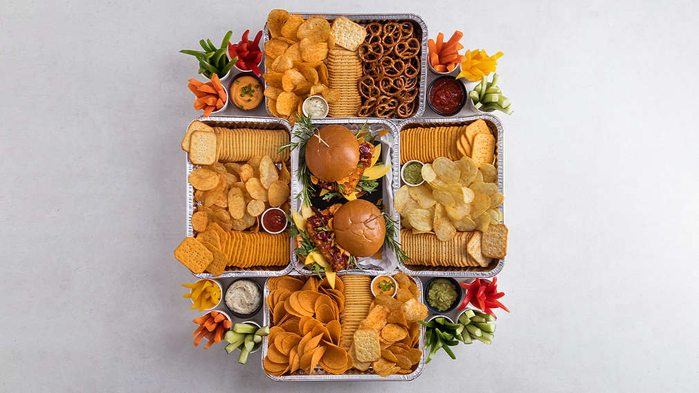 Aus der Vogelperspektive ist ein Snackstadion zu sehen, das aus mehreren Grillschalen besteht, das in der Mitte mit Guacamole, außerdem mit Gemüsesticks, Crackern und Chicken Wings befüllt ist. Rund um das Stadion sind außerdem Wraps, Sandwiches und Dips platziert.