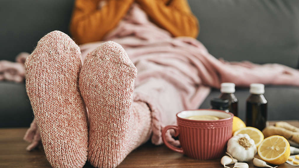 Zu sehen ist eine Person, die auf dem Sofa sitzt und ihre Füße auf dem Tisch aufgelegt hat. Auf dem Tisch sieht man eine Tasse, Knoblauch, aufgeschnittene Zitronen, Tabletten und kleine Medizinflaschen.