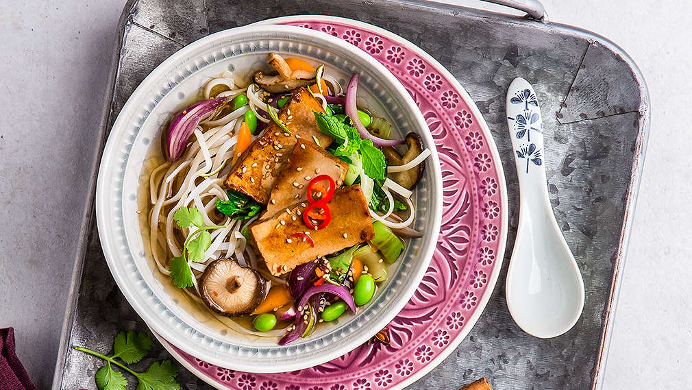 asiatische Suppenschüssel auf einem grauen Emailgeschirr, gefüllt mit einer Pho-Suppe mit Pak Choi, Nudeln und Tofuscheiben. Auf dem Tablett sind außerdem ein weinrotes Geschirrtuch, eine aufgeschnittene Limette und zwei Zimtstangen