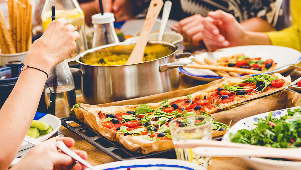 Vier Menschen im Anschnitt sitzen an einem gedeckten Tisch mit unterschiedlichen Gerichten und essen