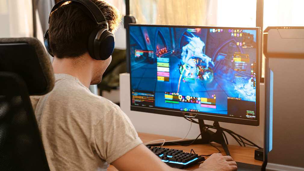 Zu sehen ist ein junger Mann, der vor dem PC sitzt und auf einem großen Bildschirm ein Computerspiel spielt