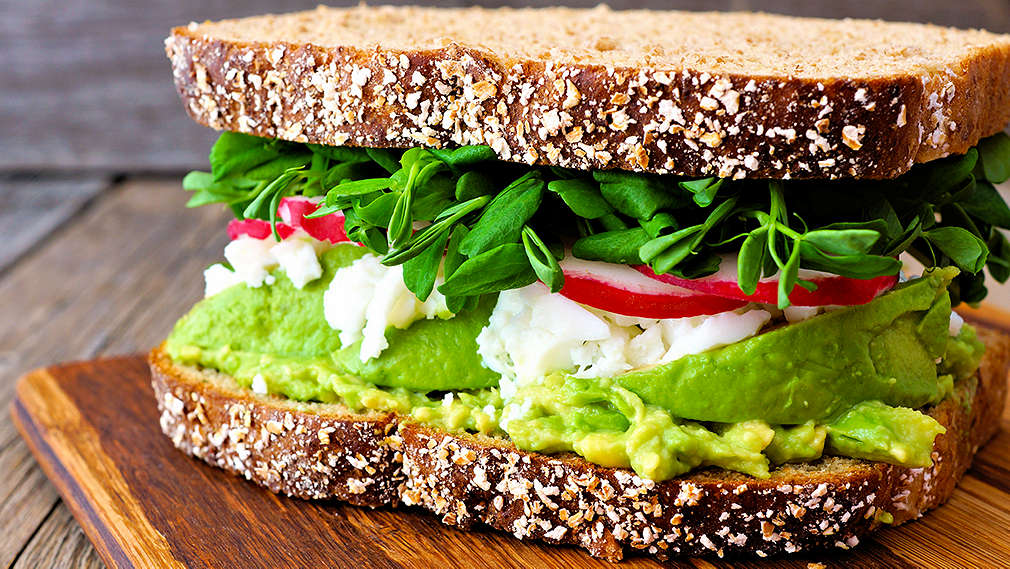 Ein mit Salat, Tomaten und Frischkäse belegtes Brot liegt auf einem Schneidebrett