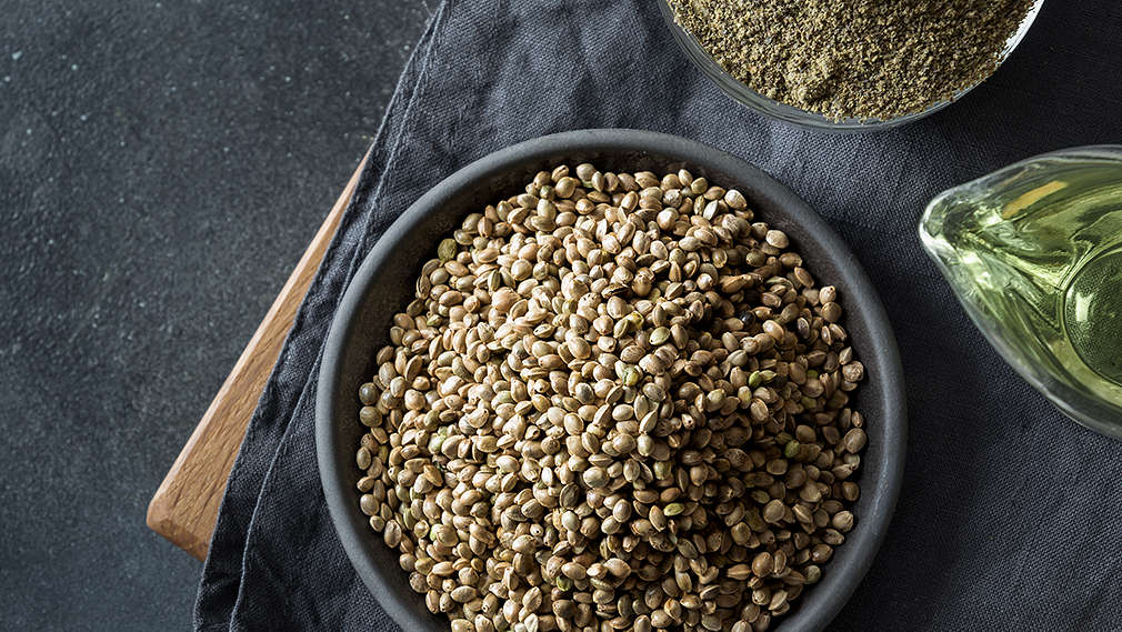 Dunkelgrauer Untergrund, darauf ein dunkelgraues Geschirrtuch, das auf einem Holzbrett liegt, das nur ganz leicht hervorlugt. Auf dem Tuch: Zwei Schalen mit je ganzen und gemahlenen Hanfsamen, außerdem ein Holzkochlöffel und eine kleine Karaffe mit Hanföl.