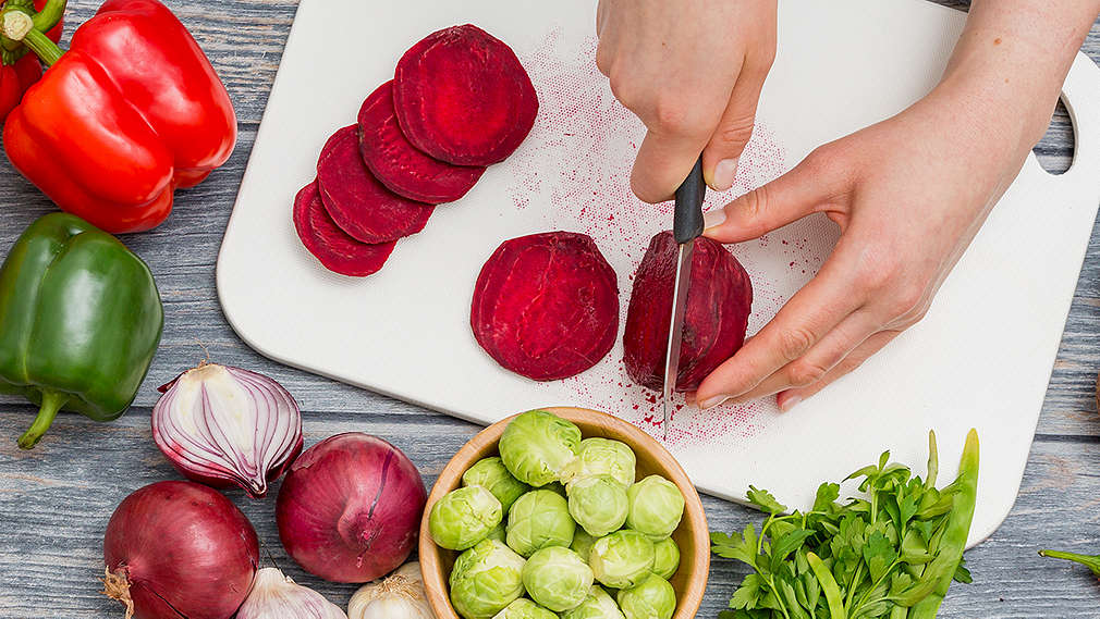 Zwei Hände die rote Beete auf einem Brett schneiden, daneben liegen Zwiebeln, Paprika, Kräuter und Rosenkohl in einer Schüssel