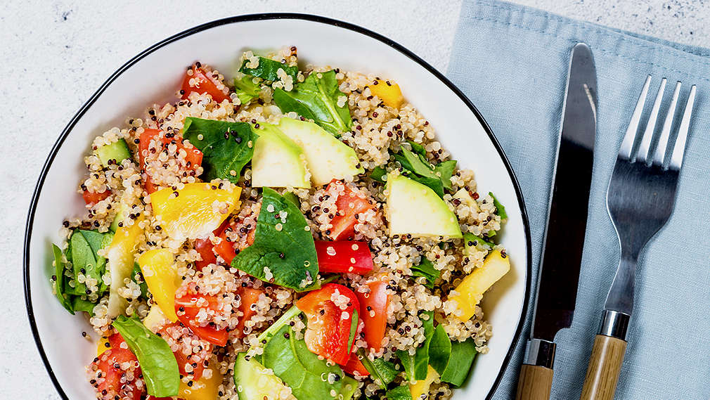 Zu sehen ist eine weiße Schale mit blauem Rand, gefüllt mit einem Gericht aus Quinoa, Paprika, Tomaten, Spinat und Avocado. Links von der Schüssel liegt eine Gabel, rechts davon eine halbe Avocado, zwei Spinatblätter und eine Cocktailtomate. In der linken Ecke des Bildes sieht man einen Teil eines weiß-grau karierten Tischtuchs