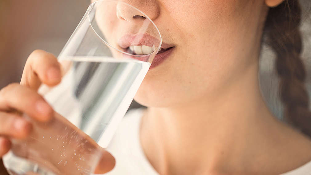 Frau, von der man im Anschnitt Gesicht und Hals sehen kann, trinkt aus einem Wasserglas