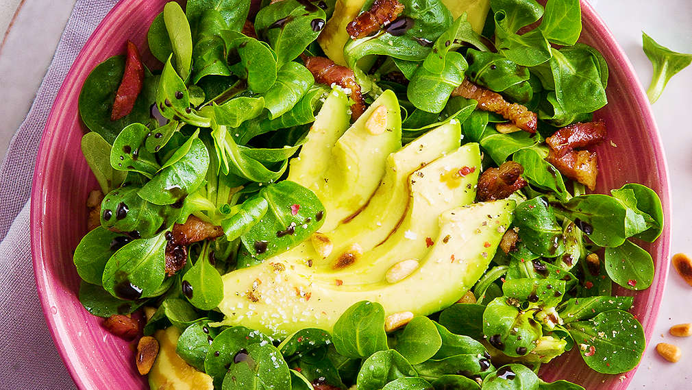 Eine von oben fotografierte pinke Schüssel, in der sich neben Feldsalat auch Avocadoscheiben, Speck und Pinienkerne befinden
