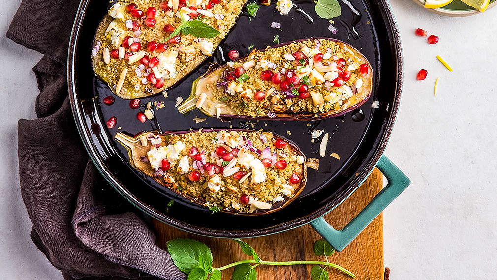 Eine Pfanne auf hellgrauem Untergrund, rechts oben ein Schälchen mit Dip, Granatapfelkernen und zwei Zitronenscheiben; unter der Pfanne liegt ein braunes Geschirrtuch, in der Pfanne liegen drei halbe Auberginen, gefüllt mit Couscous und bestreut mit Granatapfelkernen, frischer Minze und Mandelsplittern