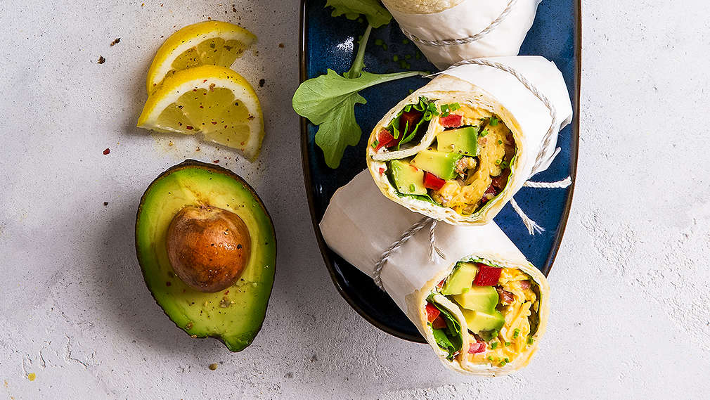 Hellgrauer Untergrund, darauf Pflücksalatblätter, Zitronenscheiben, eine halbe Avocado und ein länglicher blauer Teller mit gefüllten Wraps