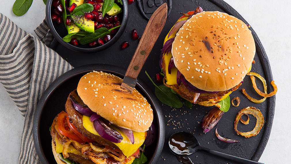 Zwei vegane Burger auf einem schwarzen, runden Teller. Die Burger sind belegt mit gegrilltem Gemüse, unter anderem Zwiebeln, Tomaten und Spinat, sowie einem veganen Burger-Patty und veganem Käse. Daneben steht in einer kleinen Schüssel ein Salat aus Avocado und Granatapfelkernen. 