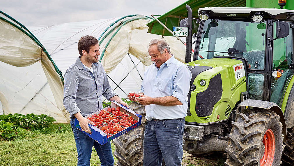 Erdbeerbauern mit Kiste Erdbeeren auf dem Feld, rechts daneben steht ein Traktor.