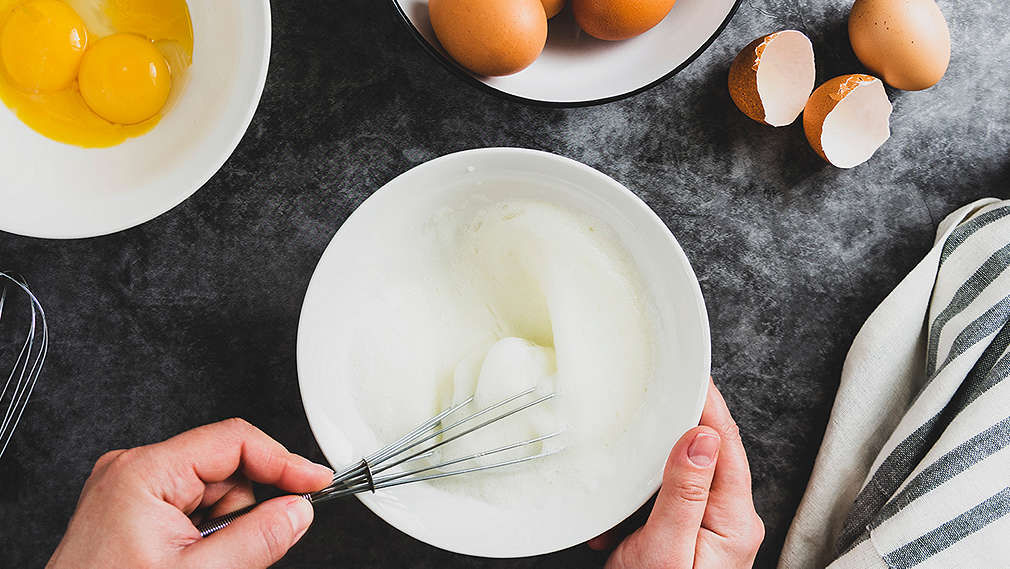 Eine Creme wird mit einem Schneebesen abgeschlagen, daneben liegen Eierschalen auf dem Küchentresen und Eigelb in einer kleinen Schüssel