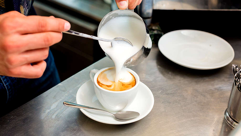 Aufgeschäumte Milch wird aus einem kleinen Kännchen mit Hilfe eines Teelöffels in eine Tasse mit Kaffee gegeben