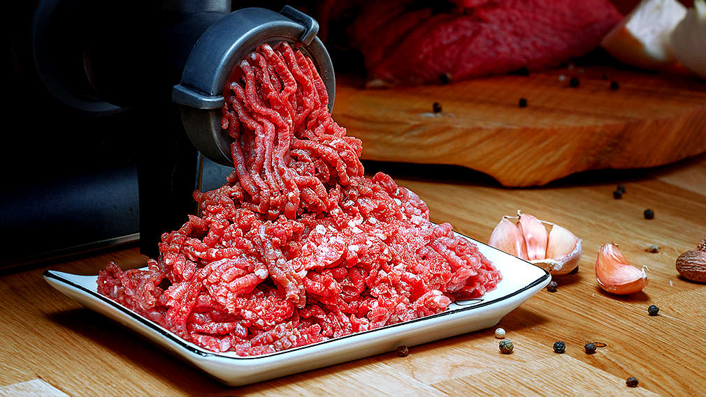 Fleisch wird durch einen Fleischwolf faschiert, daneben liegen auf einem Holztisch Pfefferkörner und Knoblauch