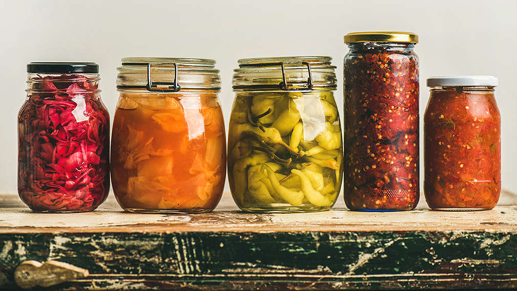 Auf einer Holzfläche stehen fünf Einmachgläser mit unterschiedlichen Sorten fermentiertem Gemüse