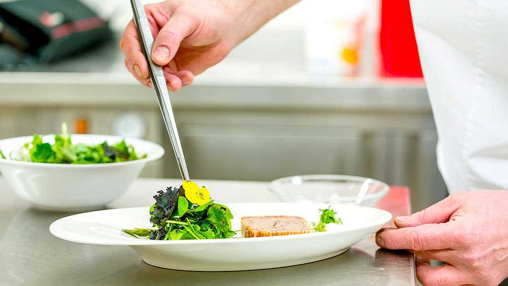 Ein weißer flacher Teller wird mit einer Pinzette garniert: Salat mit gelber Blüte und Pastete.