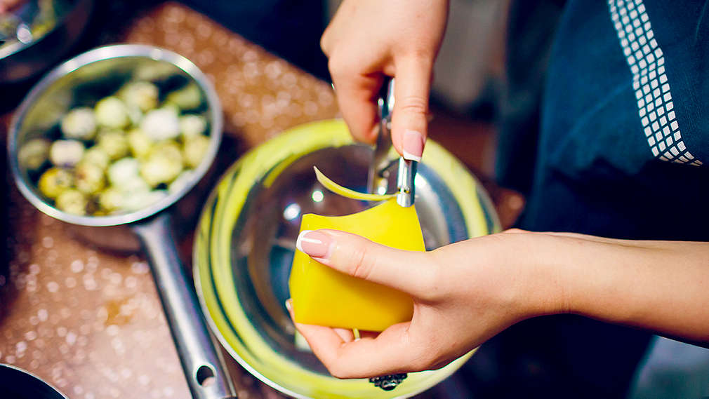 Käse wird in der Hand im Close-up in eine silberne Schüssel gehobelt. Im Hintergrund befindet sich eine Kasserolle.