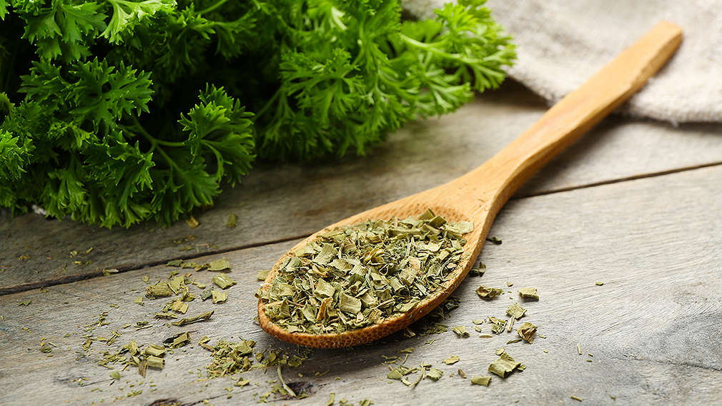 Gerebelte Petersilie in einem Holzlöffel, der auf einem Holztisch liegt. Daneben ein Bund frische Petersilie