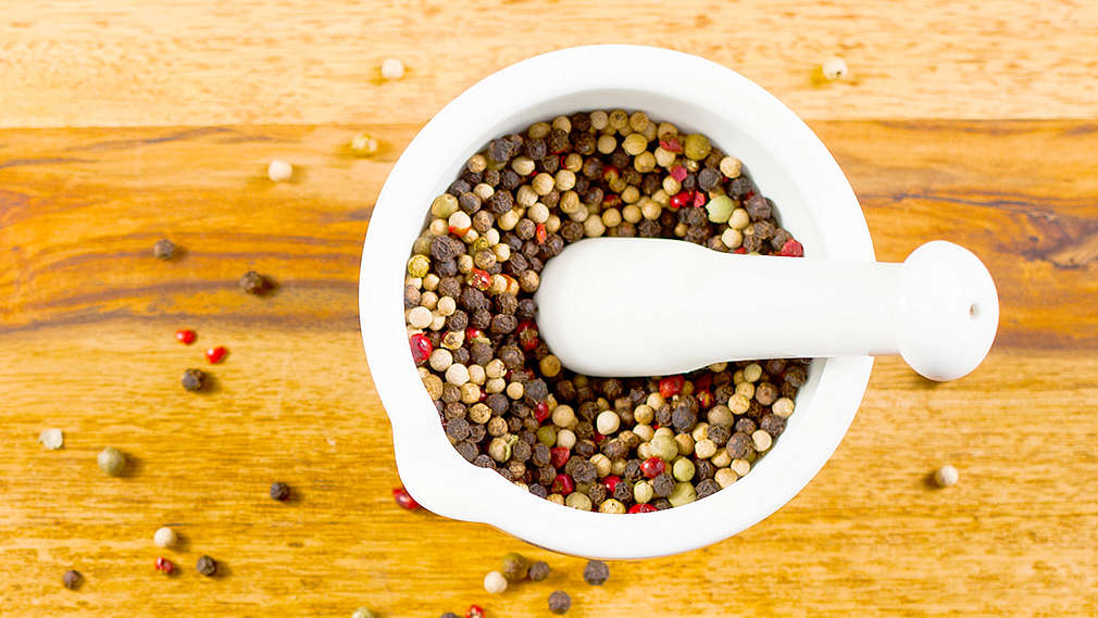 Ganze bunte Pfefferkörner in einem weißen Mörser sowie verstreut auf einem Holz-Untergrund.