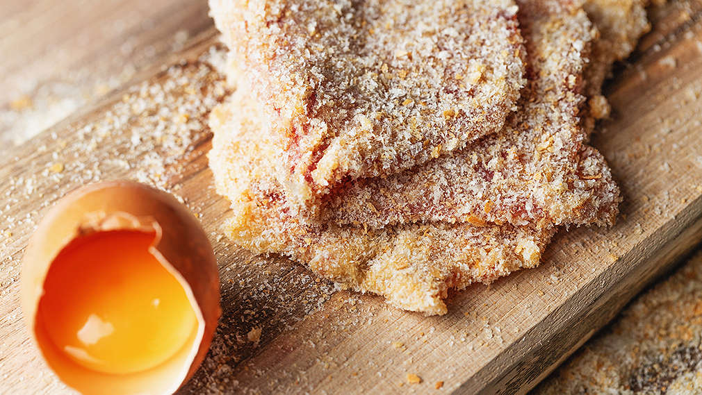 Paniertes Schnitzel liegt auf einem Holzbrett, daneben ein aufgeschlagenes Ei