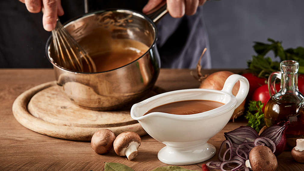 Dunkle Soße wird in einem kleinen Topf mit einem Schneebesen umgerührt, daneben steht eine Sauciere mit dunkler Soße sowie unterschiedliche Zutaten wie Champignons, Zwiebeln, Tomaten, Gewürze und ein Kännchen mit Öl