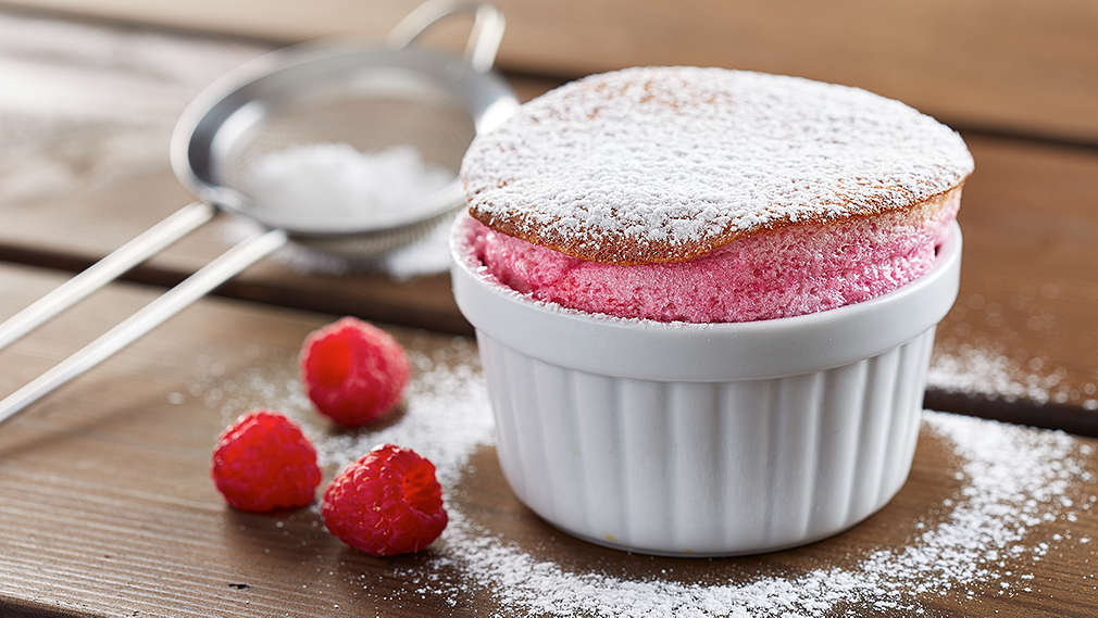 Himbeersoufflé in einer kleinen weißen Backform mit Puderzucker garniert, daneben frische Himbeeren