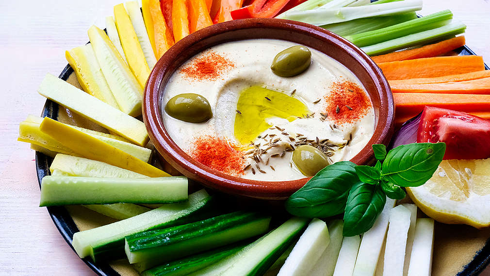 Verschiedenes Gemüse in Stiften geschnitten und auf einem Teller fecherförmig ausgelegt; in der Mitte eine Schale mit Dip, Gewürzen und Oliven