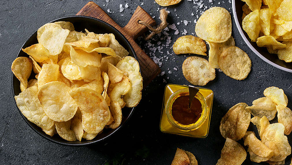 Selbstgemachte Kartoffelchips auf einem dunklen Grund sowie in zwei Schälchen