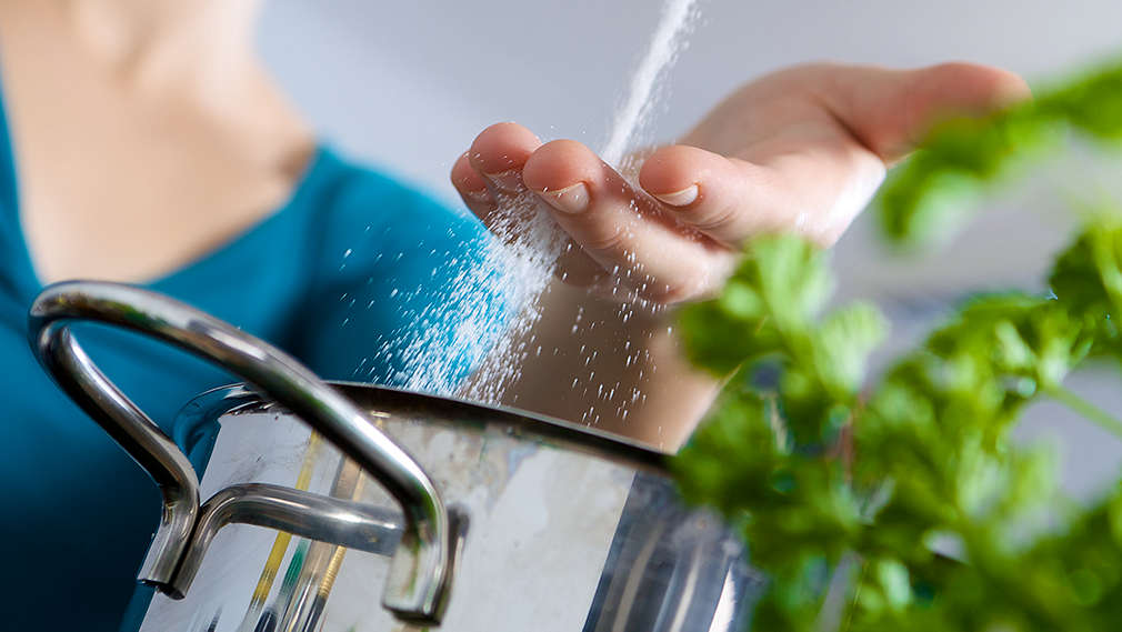 Person salzt Essen in einem Topf, daneben steht ein Töpfchen mit Basilikum