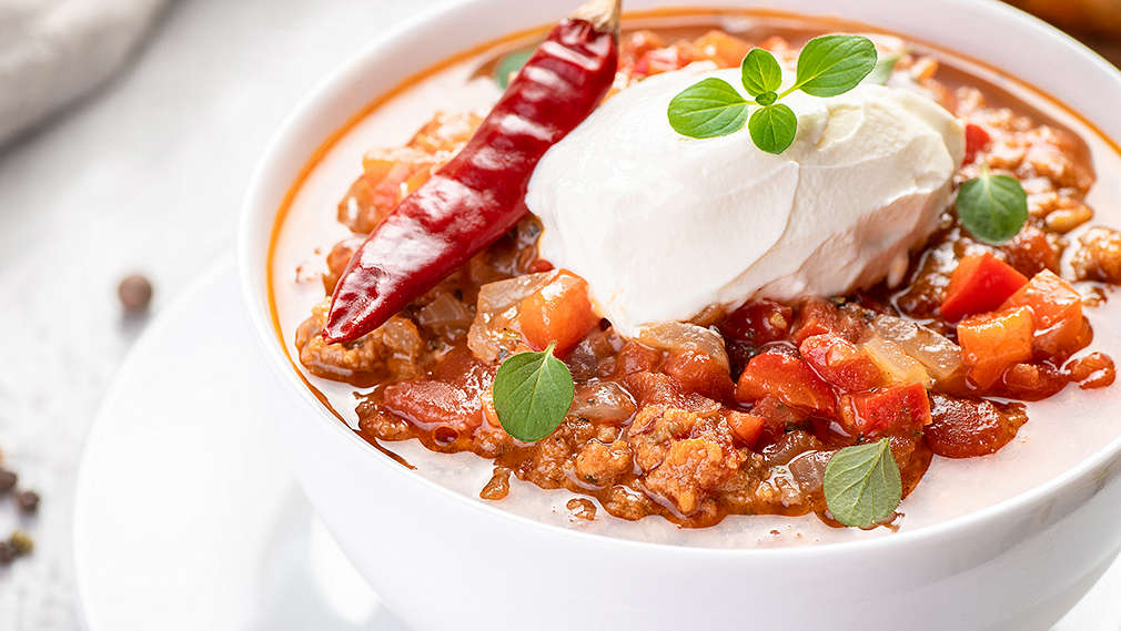 Ein Teller mit Chili con Carne mit einem Klecks Schmand obendrauf. Ebenfalls darauf liegt eine Chilischote und frische Kräuter. 