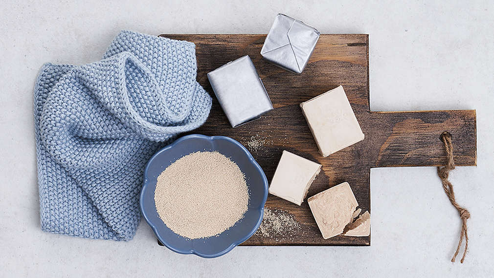 Zu sehen ist ein teilweise zerbröckelter Hefewürfel auf dunkler Arbeitsfläche, im Hintergrund ein ganzer Hefewürfel.