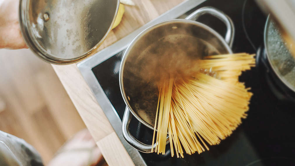Ein Topf auf dem Herd mit kochendem Wasser und Spaghetti; der Deckel wird angehoben