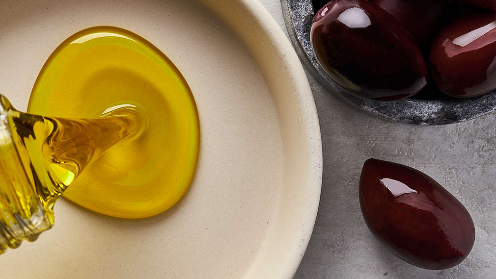 Heller Untergrund, auf dem ein helles Schälchen zu sehen ist, in das Öl aus einer Flasche gegossen wird, daneben ein kleineres dunkelgraues Schälchen mit ganzen schwarzen Oliven.