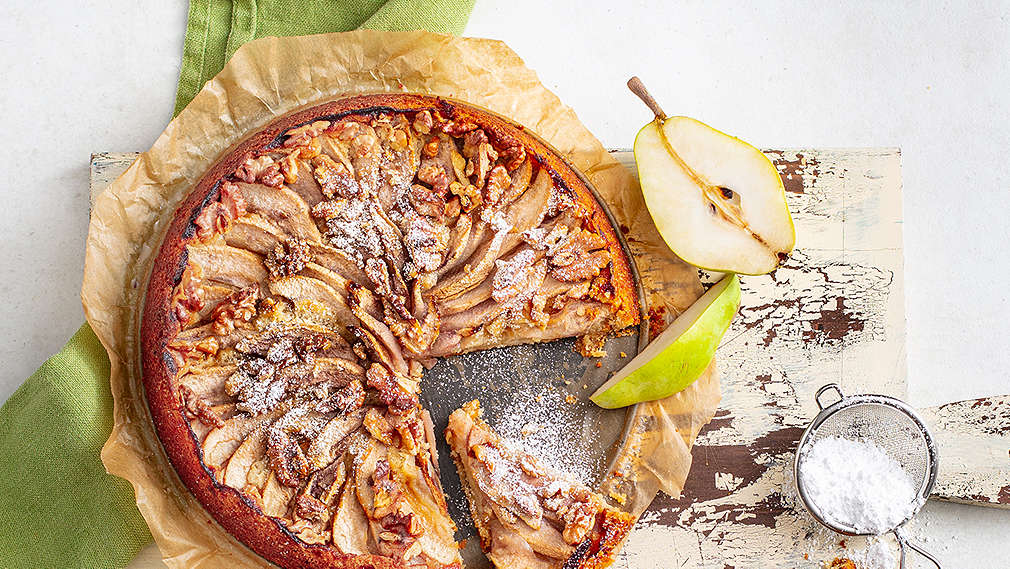 Grauer Untergrund, darauf ein hell lackiertes Holzbrettchen und ein grünes Geschirrtuch, darauf eine runde Backform mit angeschnittenem Kuchen, der mit Birnenscheiben belegt ist. Außerdem im Bild: Halbierte Birne, Kuchenstück, kleines Sieb mit Puderzucker, Schälchen mit Apfelmus.