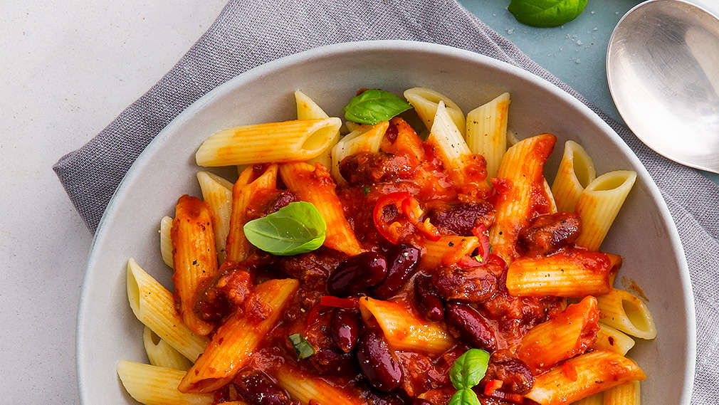 Ein tiefer hellgrauer Teller steht auf hellgrauem Untergrund und einem grauen Geschirrtuch, darin befindet sich eine große Portion Penne mit roter Soße aus Tomaten, Chili und Kindeybohnen, als Garnitur sind einige Blättchen Basilikum auf den Nudeln.