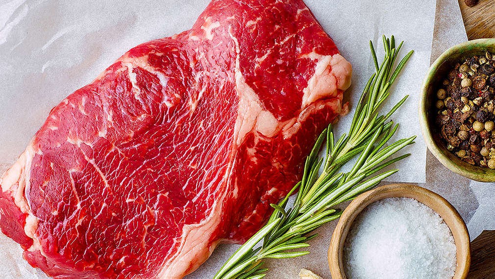 Auf einem Tisch liegt ein rohes Steak auf weißem Papier. Daneben liegt ein Zweig Rosmarin und es stehen zwei kleine Schälchen mit Salz und Pfeffer daneben
