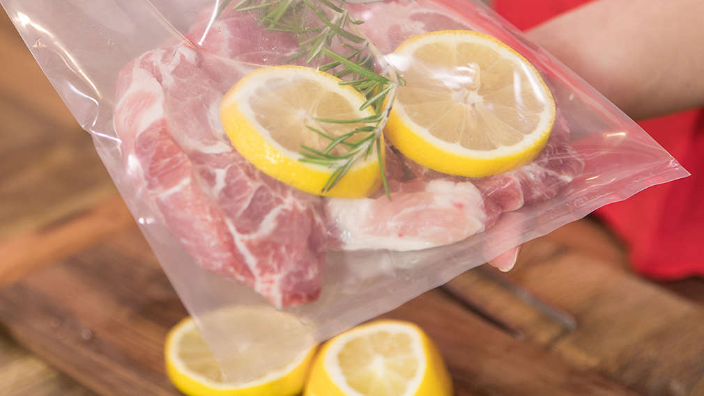 Oberkörper einer Frau im Anschnitt, zwei Hände halten einen Plastikbeutel, darin befindet sich ein Stück Fleisch, Zitronenscheiben und ein Zweig Kräuter. Die Frau arbeitet über einer Arbeitsfläche aus dunklem Holz. 