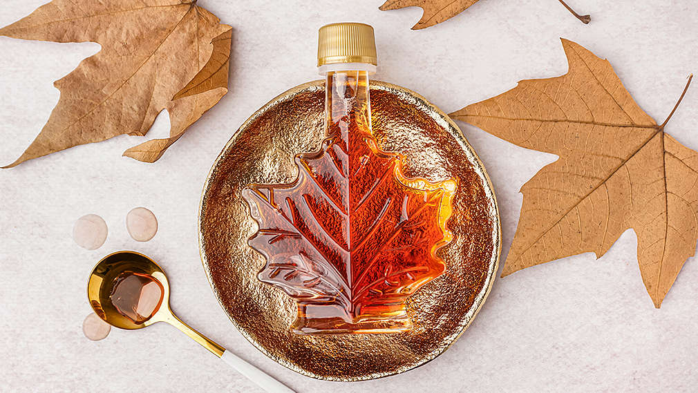 Eine Flasche Ahornsirup liegt auf einem goldenen Teller, daneben ein goldfarbener Löffel mit Ahornsirup sowie einige Ahornblätter