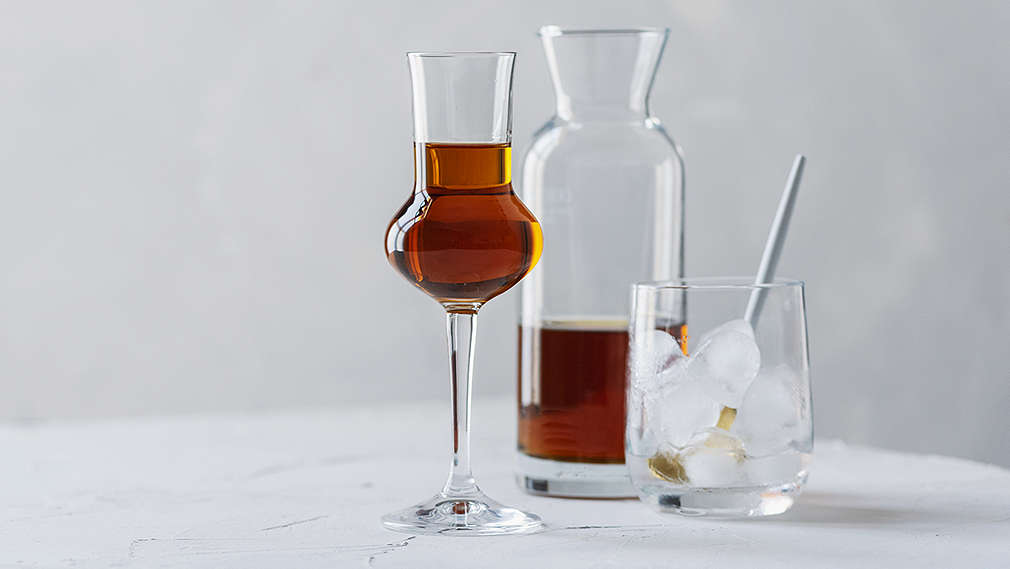 Zu sehen ist ein hohes Schnapsglas mir brauner Flüssigkeit, daneben eine gefüllte Karaffe und ein Glas mit Eiswürfeln