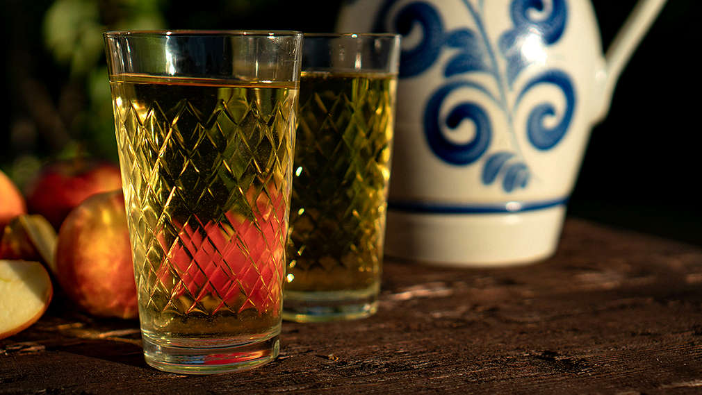 Zwei Gläser Apfelwein, sogenannte Gerippte, auf einem Holztisch, daneben ein Bembel sowie Äpfel im Hintergrund