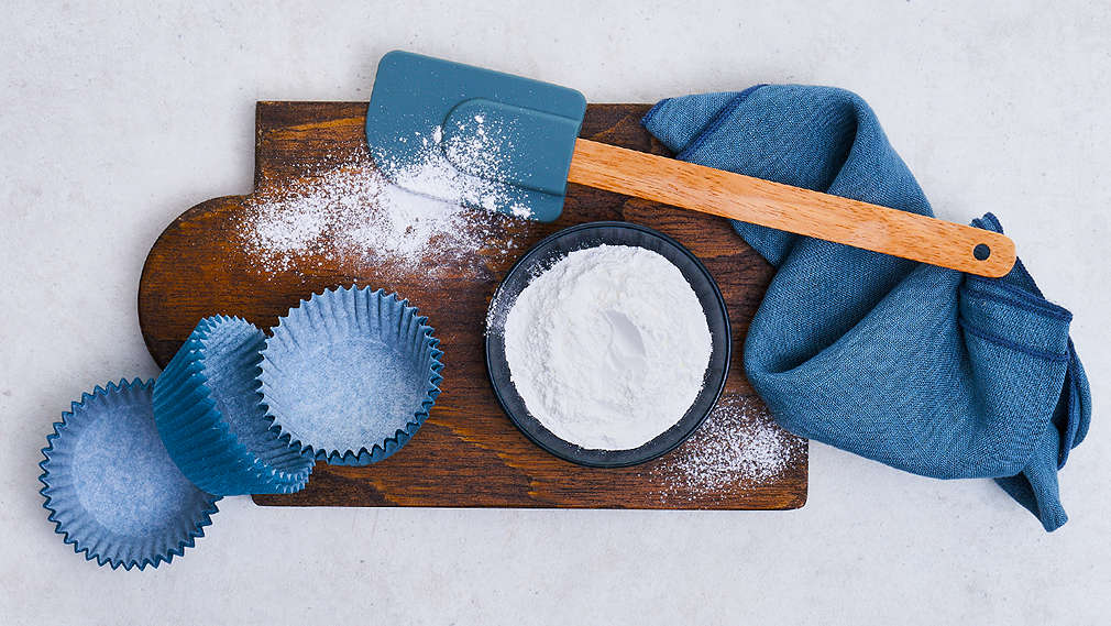 Schälchen Backpulver, Teigschaber und Muffinförmchen auf einem dunklen Holzbrettchen