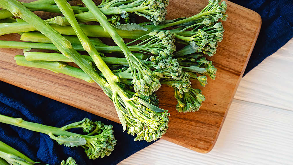 Mehrere Stiele Bimi liegen mittig auf einem Schneidebrett auf weißem Untergrund aus Holz sowie links daneben auf einer dunkel blauen Tischdecke