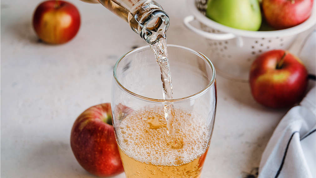 Cidre wird in ein Glas geleert, daneben liegen Äpfel auf dem Tisch sowie in einem weißen Sieb