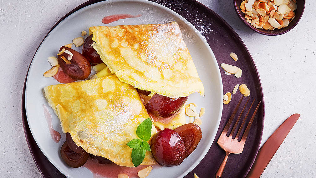 Zwei zusammengefaltete, geviertelte, dünne Pfannkuchen auf einem Teller mit Sauce, eingekochten Pflaumen, Zitronenmelisse und Mandelblättchen serviert