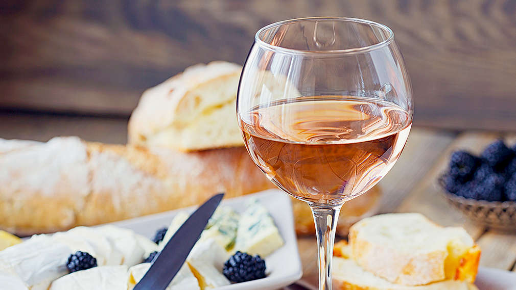 Ein Glas Dessertwein, serviert zu Käse und Baguette, welches teilweise in Scheiben geschnitten ist. Im Hintergrund ist eine kleine Schale mit Blaubeeren garniert.