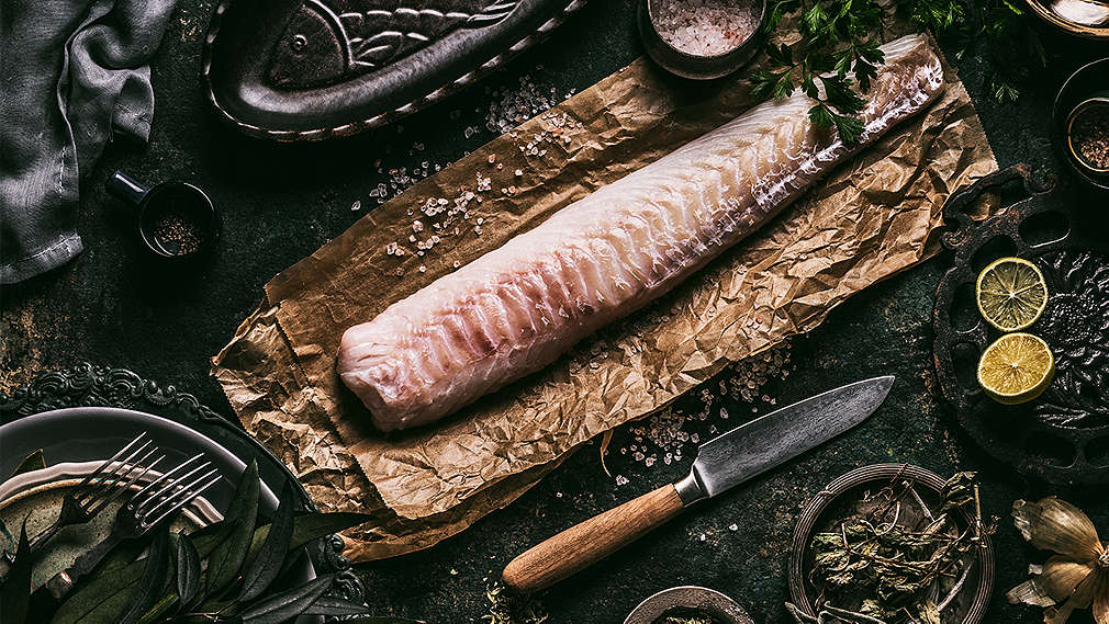 Zu sehen ist Dorschfilet auf Backpapier, daneben Küchenutensilien 