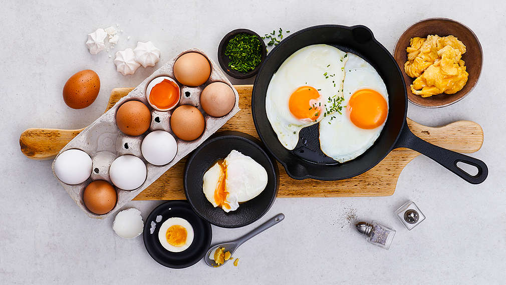 Auf einem Holzschneidebrett auf einem Esstisch stehen Eier in verschiedener Form: rohe Eier in einer Verpackung, Spiegelei in einer Bratpfanne, pocherte Eier in einerm Teller und Omelett in einer kleinen Schale. Daneben steht eine kleine Schale mit geschnittenem Lauch und auf der anderen Seite liegt einen Salz- und Pfefferstreuer. Alles wurde von oben abfotografiert.